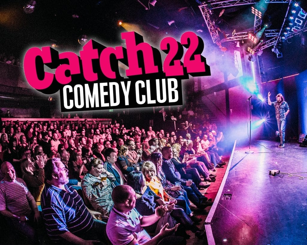 A promotional photo for Catch 22 Comedy Club. The vibrant scene depicts a crowded comedy venue with a large audience seated in front of a stage. On stage, a performer (likely a comedian) can be seen standing with a microphone. The venue is lit with theatrical lighting in purple and blue hues, creating an energetic atmosphere.