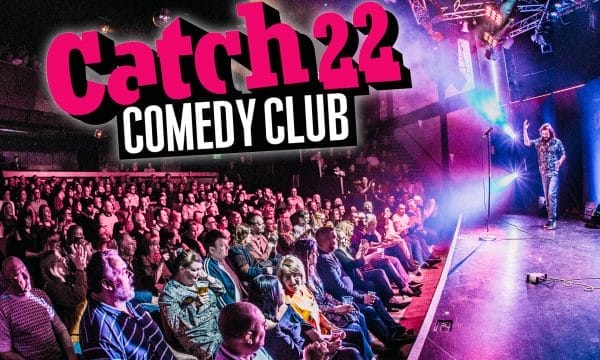 A promotional photo for Catch 22 Comedy Club. The vibrant scene depicts a crowded comedy venue with a large audience seated in front of a stage. On stage, a performer (likely a comedian) can be seen standing with a microphone. The venue is lit with theatrical lighting in purple and blue hues, creating an energetic atmosphere.