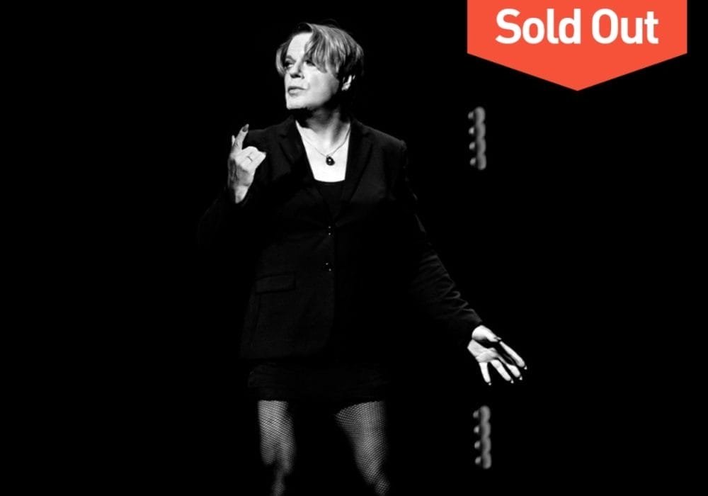 Comedian Eddie Izzard, a white person with medium length hair, stands against a black backdrop. They are wearing a black jacket, and a black necklace. The image is black and white.
