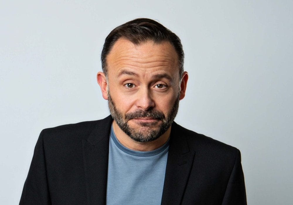 Geoff Norcott, a white middle aged man with short brown hair and a short beard, looks directly into the camera with a slight smirk. He is wearing a black suit jacket, with a blue tshirt.