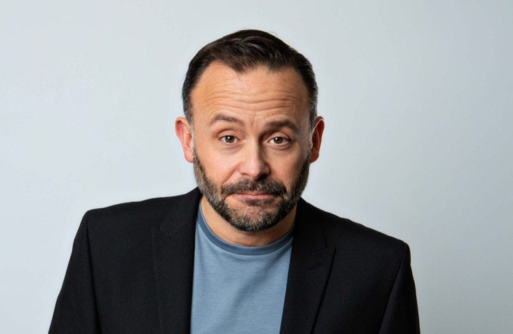 Geoff Norcott, a white middle aged man with short brown hair and a short beard, looks directly into the camera with a slight smirk. He is wearing a black suit jacket, with a blue tshirt.