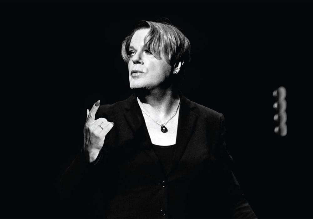 Comedian Eddie Izzard, a white person with medium length hair, stands against a black backdrop. They are wearing a black jacket, and a black necklace. The image is black and white.