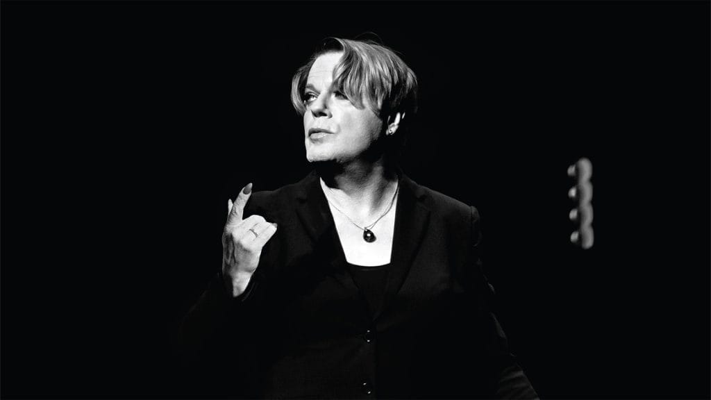 Comedian Eddie Izzard, a white person with medium length hair, stands against a black backdrop. They are wearing a black jacket, and a black necklace. The image is black and white.