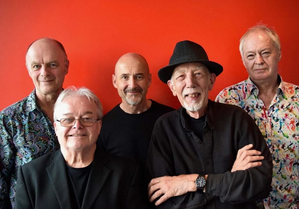 I can see an image of five older men standing together against a bright red background. There are three men in the back row and two men in the front. The men are dressed in various outfits - some wearing patterned shirts while others are in black clothing. One man in the front row is wearing a black hat. They appear to be posing for a group photograph.
