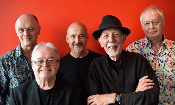 I can see an image of five older men standing together against a bright red background. There are three men in the back row and two men in the front. The men are dressed in various outfits - some wearing patterned shirts while others are in black clothing. One man in the front row is wearing a black hat. They appear to be posing for a group photograph.