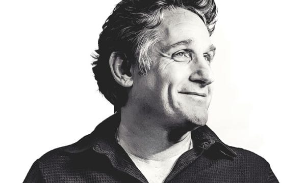Tom stands with wavy, greying hair and a slight smile, looking off to the side. He is wearing what appears to be a dark button-up shirt or polo. The photograph has a professional, artistic quality with good contrast and lighting that highlights his facial features.