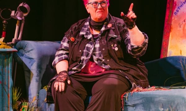 a person with bright pink hair seated on what appears to be a blue couch or chair in what looks like a stage setting. They're wearing glasses, a plaid shirt over dark clothing, and lace gloves or wristbands. The person is gesturing with one hand raised, appearing to be speaking or performing. The setting has a dark background with some colourful elements visible to the side, including what looks like decorative items and possibly a screen or backdrop with vibrant colours.