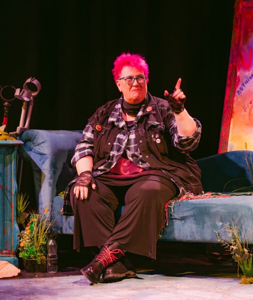 a person with bright pink hair seated on what appears to be a blue couch or chair in what looks like a stage setting. They're wearing glasses, a plaid shirt over dark clothing, and lace gloves or wristbands. The person is gesturing with one hand raised, appearing to be speaking or performing. The setting has a dark background with some colourful elements visible to the side, including what looks like decorative items and possibly a screen or backdrop with vibrant colours.