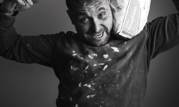 a black and white image of a white man in his mid-thirties grinning. He is wearing a black jumper covered in white paint and holding a bag of plaster on his left shoulder.