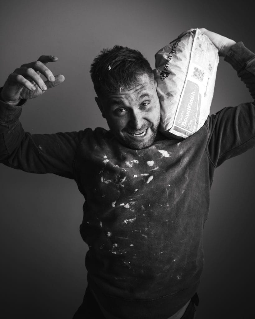 a black and white image of a white man in his mid-thirties grinning. He is wearing a black jumper covered in white paint and holding a bag of plaster on his left shoulder.