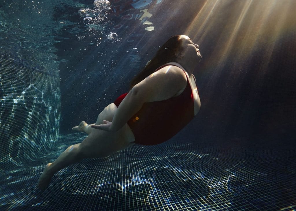 Woman in red swimsuit swimming underwater in a pool with sunbeams.