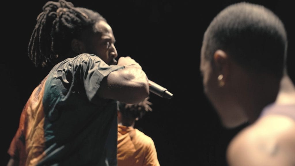 Two black men on a dark background. One is holding a microphone and wearing a black, orange and green t-shirt and has dreadlocks. The man in the foreground is topless and has a shaved head.