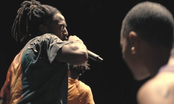 Two black men on a dark background. One is holding a microphone and wearing a black, orange and green t-shirt and has dreadlocks. The man in the foreground is topless and has a shaved head.