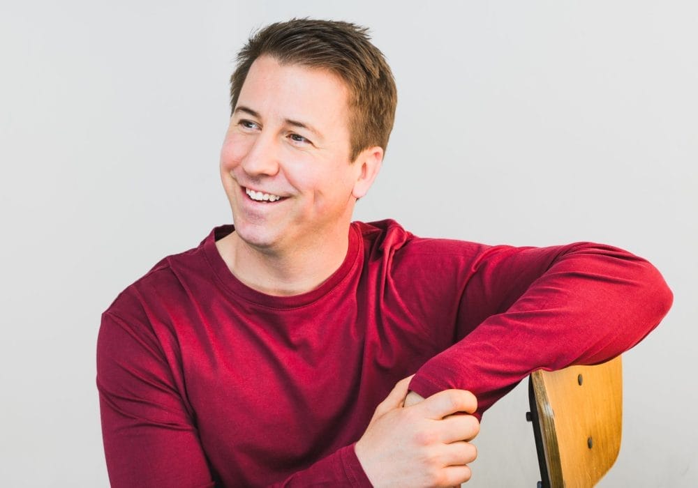 A man is wearing a bright red shirt with his arm resting on the back of a chair, he is looking off to the left and smiling. the background is grey.