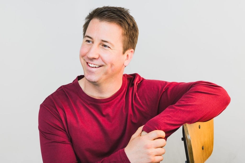 A man is wearing a bright red shirt with his arm resting on the back of a chair, he is looking off to the left and smiling. the background is grey.