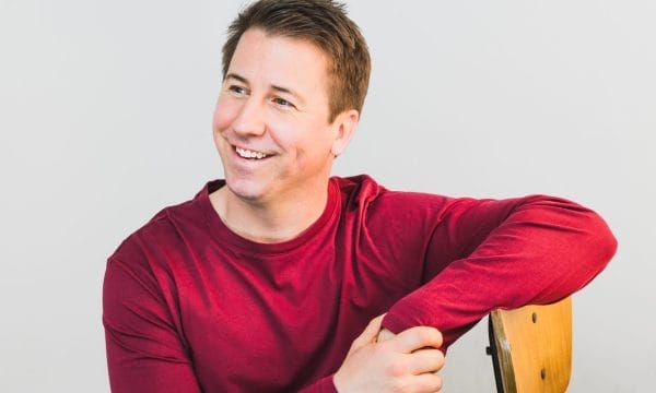 A man is wearing a bright red shirt with his arm resting on the back of a chair, he is looking off to the left and smiling. the background is grey.