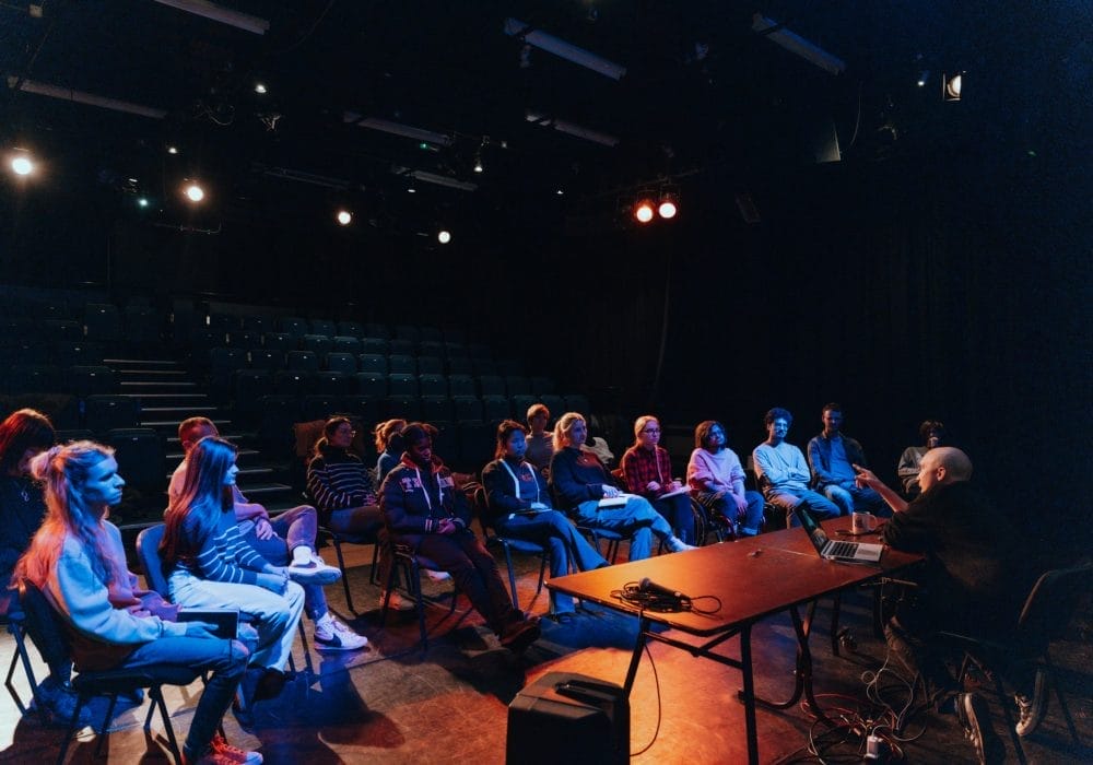 A dark image in a theatre, seating can be seen in the back and there is a group of people sat down around a man who is sat behind a table with a computer, talking to them.