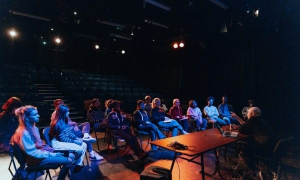 A dark image in a theatre, seating can be seen in the back and there is a group of people sat down around a man who is sat behind a table with a computer, talking to them.