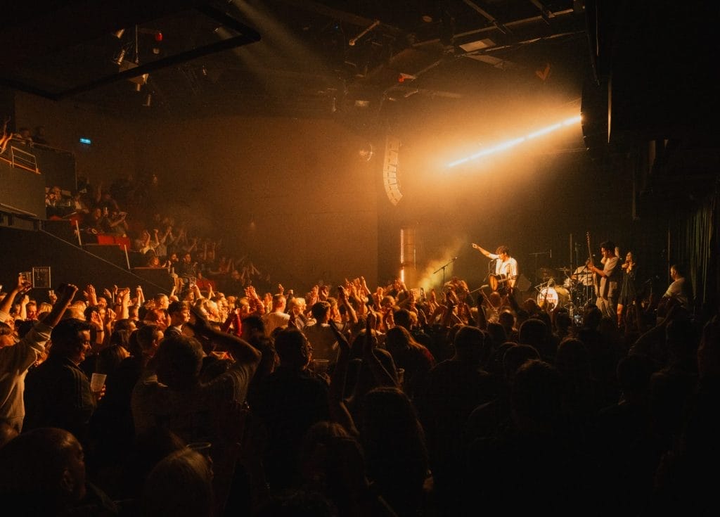 A lively concert scene inside a packed venue, with a large crowd cheering and raising their hands toward the stage. Warm stage lighting illuminates the performer, who is pointing into the audience while playing guitar, backed by a full band.