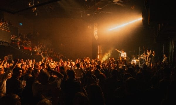 A lively concert scene inside a packed venue, with a large crowd cheering and raising their hands toward the stage. Warm stage lighting illuminates the performer, who is pointing into the audience while playing guitar, backed by a full band.