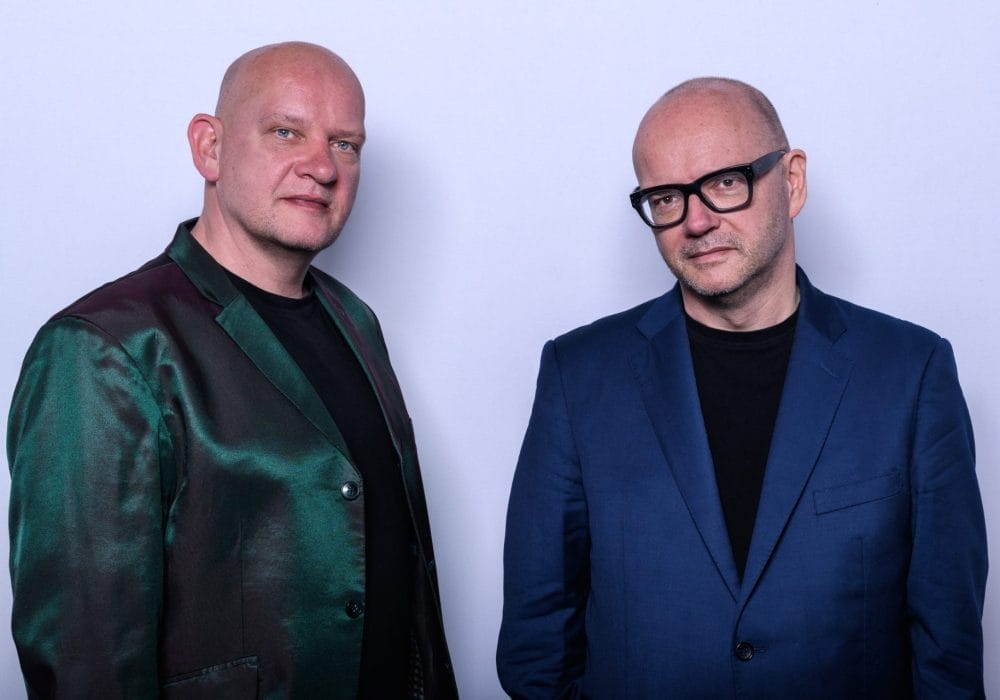 Two men stand side by side against a plain light background. Both are bald and wearing black shirts; the man on the left wears a shiny green and burgundy jacket, while the man on the right wears a blue suit jacket and black-rimmed glasses. They look calmly into the camera.