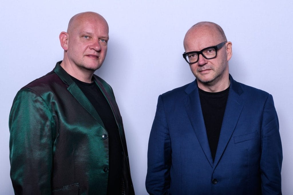 Two men stand side by side against a plain light background. Both are bald and wearing black shirts; the man on the left wears a shiny green and burgundy jacket, while the man on the right wears a blue suit jacket and black-rimmed glasses. They look calmly into the camera.