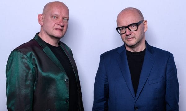 Two men stand side by side against a plain light background. Both are bald and wearing black shirts; the man on the left wears a shiny green and burgundy jacket, while the man on the right wears a blue suit jacket and black-rimmed glasses. They look calmly into the camera.