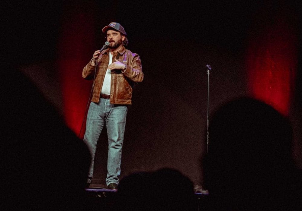 Man in a brown leather jacket stands on stage, holding a microphone, performing comedy in front of a dark audience.