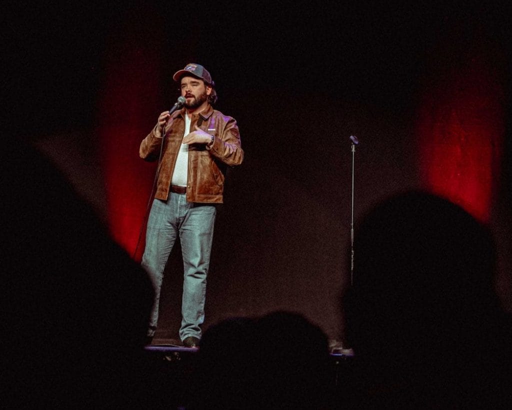 Man in a brown leather jacket stands on stage, holding a microphone, performing comedy in front of a dark audience.