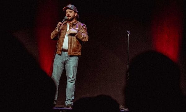 Man in a brown leather jacket stands on stage, holding a microphone, performing comedy in front of a dark audience.
