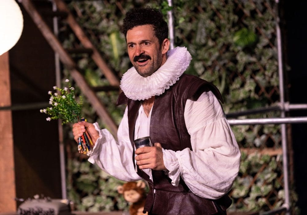 Man in Elizabethan costume, holding flowers and pencils, on a stage.
