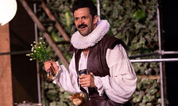 Man in Elizabethan costume, holding flowers and pencils, on a stage.
