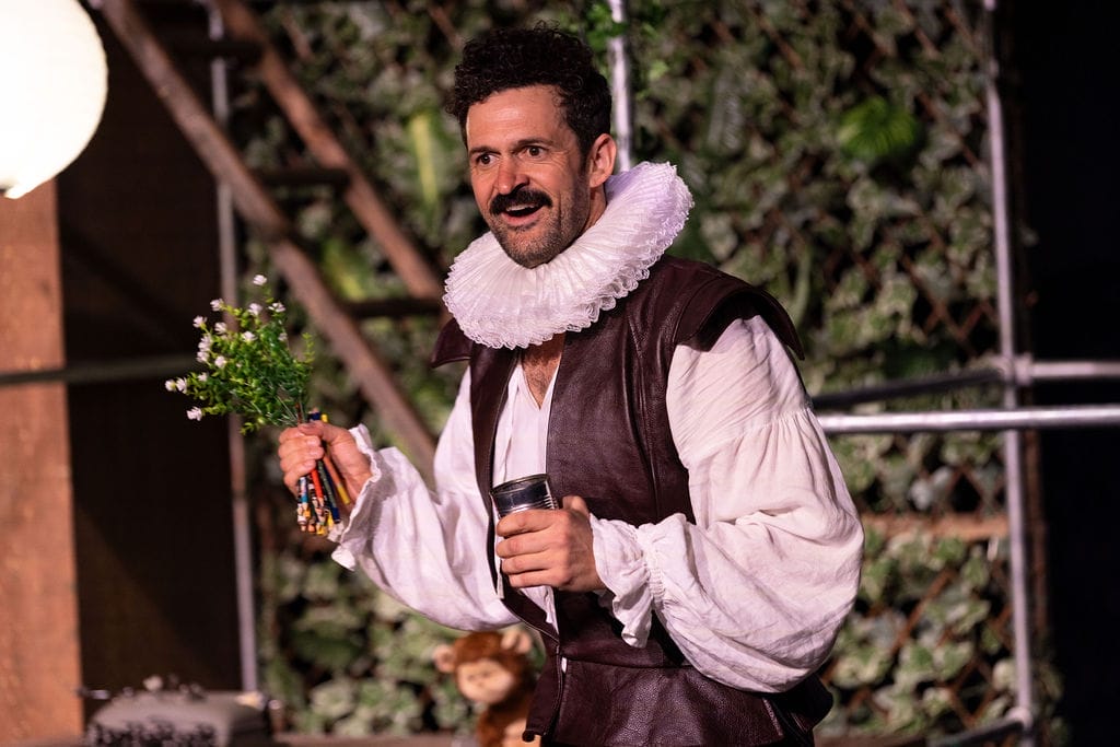 Man in Elizabethan costume, holding flowers and pencils, on a stage.