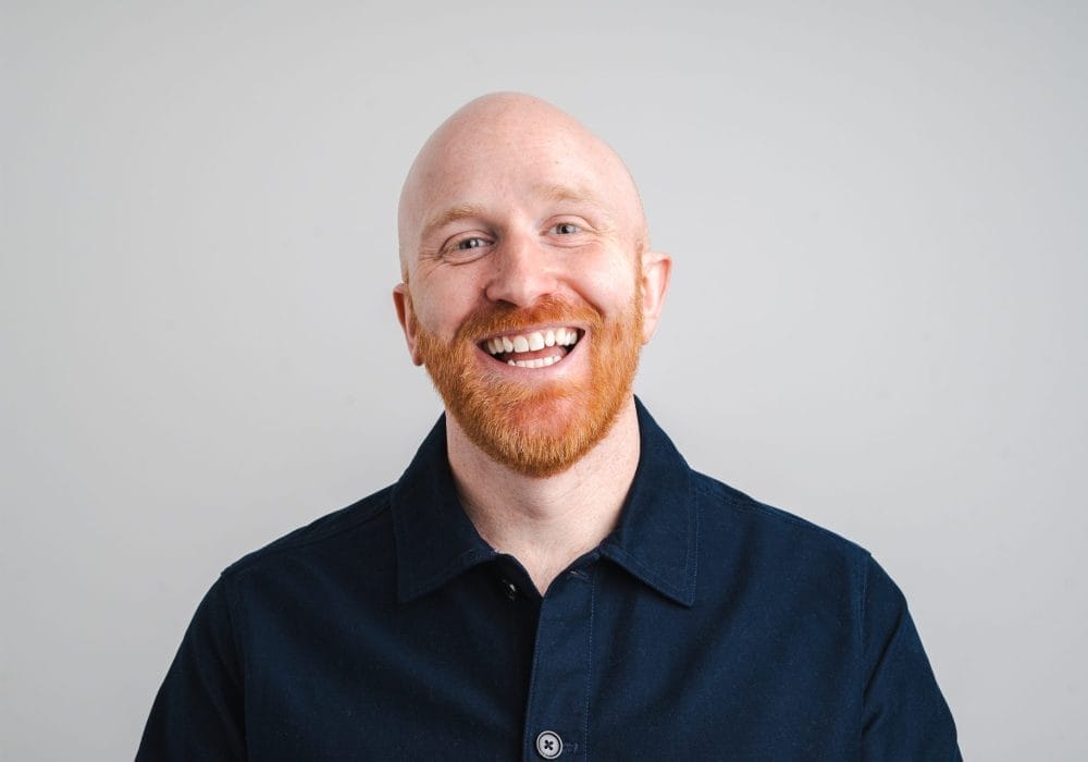 A man smiling, wearing a blue shirt on a grey background.