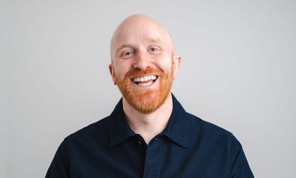 A man smiling, wearing a blue shirt on a grey background.