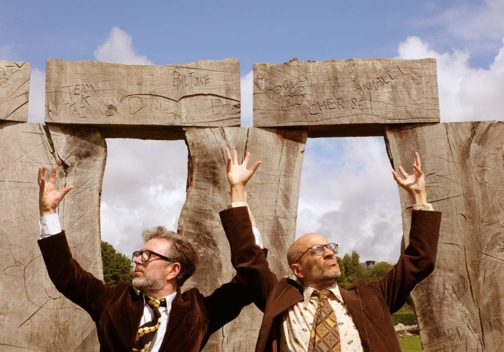 Two men in brown jackets and patterned ties, arms raised, in front of large, carved wooden pillars.