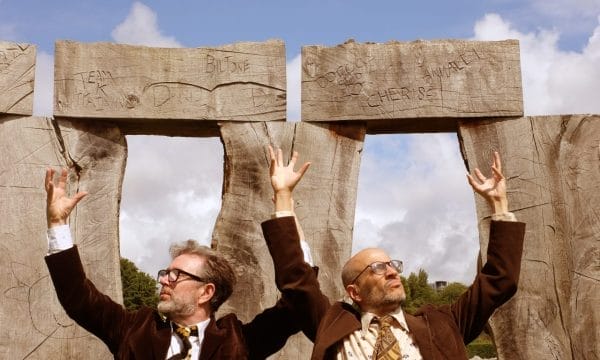 Two men in brown jackets and patterned ties, arms raised, in front of large, carved wooden pillars.