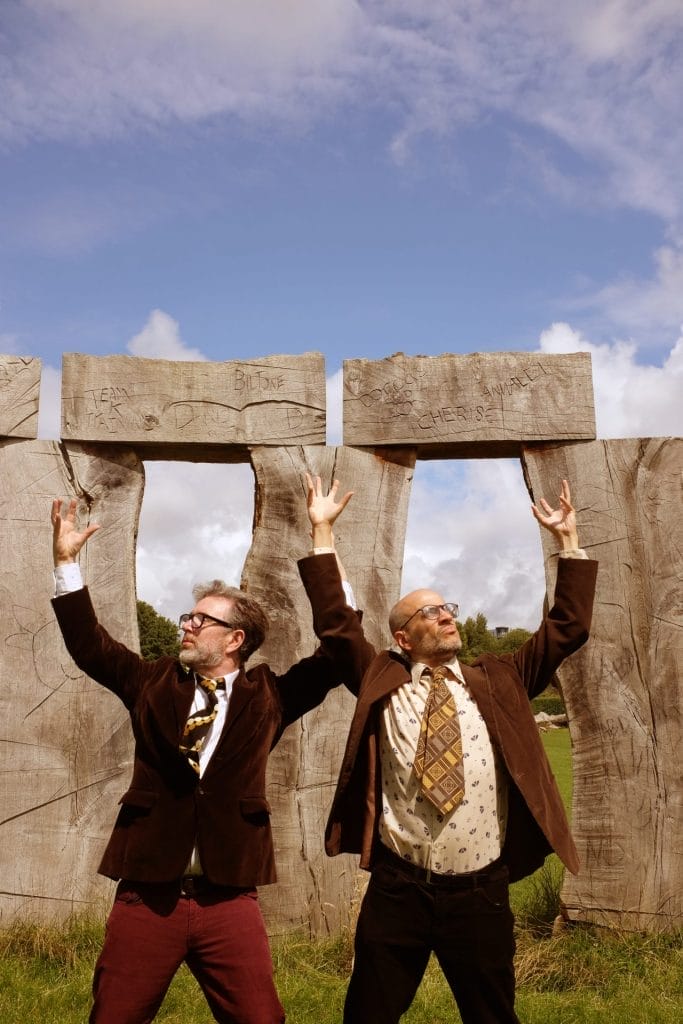 Two men in brown jackets and patterned ties, arms raised, in front of large, carved wooden pillars.