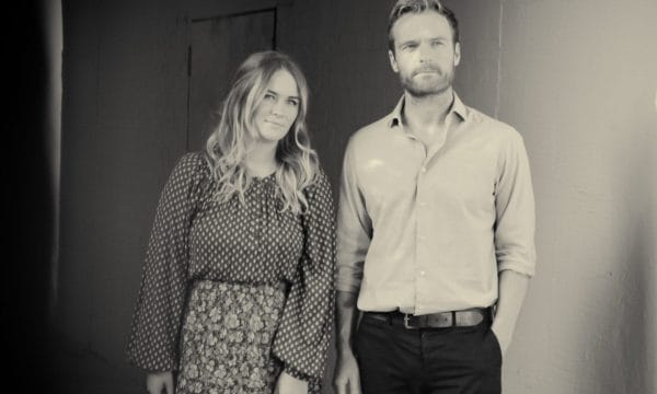 two people standing side by side against a wall in a black and white photograph. On the left is a person wearing a patterned dress with mixed prints that appears to have a flowing, tiered design. On the right is a person in a light-coloured button-up shirt and dark trousers with a belt. There's interesting lighting in the scene, with what looks like dappled light or shadows falling on the wall to the right side of the frame. The photograph has a vintage or artistic quality due to the monochrome treatment and the composition.