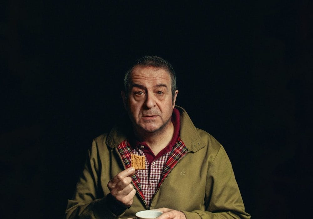 A black background with an older middle-aged man in a tan jacket, holding a biscuit and a coffee cup, looking directly at the camera.