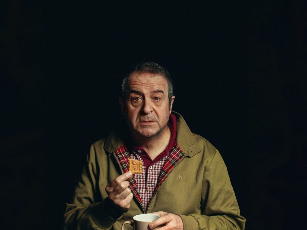 A black background with an older middle-aged man in a tan jacket, holding a biscuit and a coffee cup, looking directly at the camera.