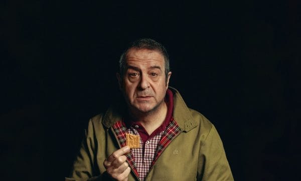 A black background with an older middle-aged man in a tan jacket, holding a biscuit and a coffee cup, looking directly at the camera.