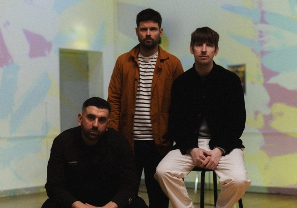 An image of three men, the one on the left is sitting on a yellow chair, wearing all black, there is a man stood up behind him wearing a stripy shirt and an orange unbuttoned shirt, the man to his right is on a stool wearing a black shirt and white trousers.
