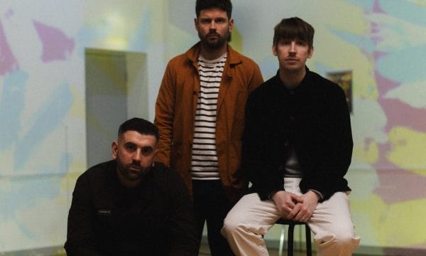 An image of three men, the one on the left is sitting on a yellow chair, wearing all black, there is a man stood up behind him wearing a stripy shirt and an orange unbuttoned shirt, the man to his right is on a stool wearing a black shirt and white trousers.
