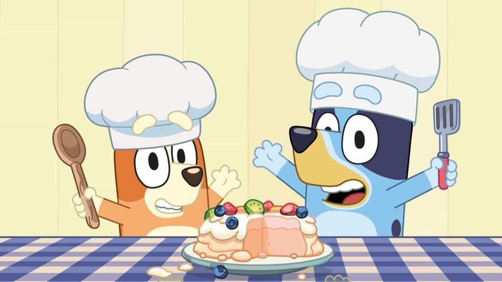 A cartoon image of two of the characters from Bluey wearing chef's hats and holding cooking utensils. There is a plate with some food on a checkered blue table cloth. The background is a pale yellow.