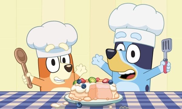 A cartoon image of two of the characters from Bluey wearing chef's hats and holding cooking utensils. There is a plate with some food on a checkered blue table cloth. The background is a pale yellow.