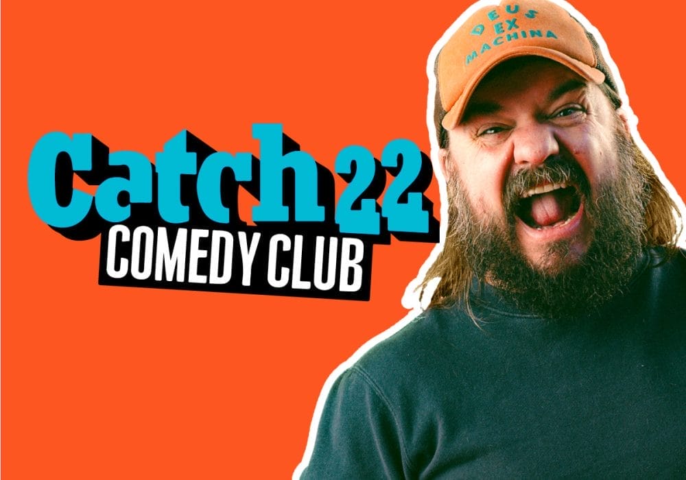 a promotional header for "Catch22 Comedy Club" with a bright orange background. The club's name appears in turquoise and white letters on the left side. On the right is a bearded man with long hair wearing an orange cap that reads "DEUS EX MACHINA" and a dark shirt. He appears to be laughing or shouting enthusiastically with his mouth open wide.