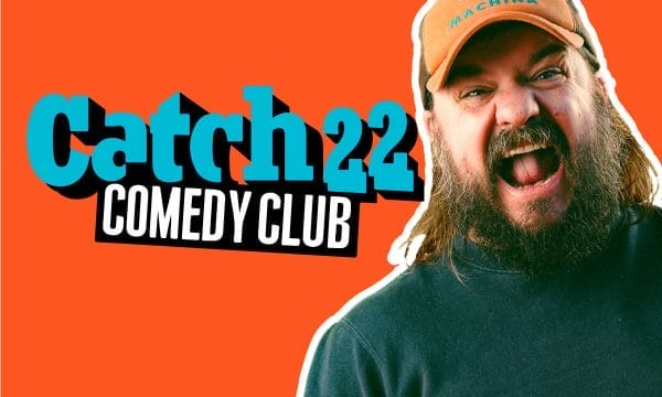 a promotional header for "Catch22 Comedy Club" with a bright orange background. The club's name appears in turquoise and white letters on the left side. On the right is a bearded man with long hair wearing an orange cap that reads "DEUS EX MACHINA" and a dark shirt. He appears to be laughing or shouting enthusiastically with his mouth open wide.