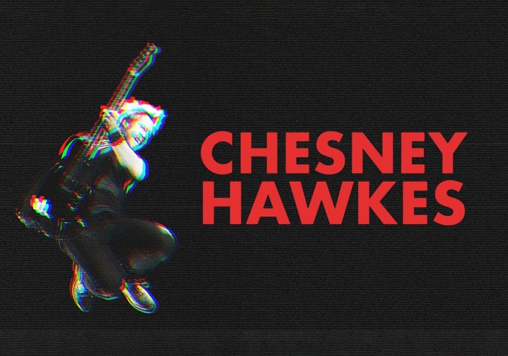 An image with red writing that reads 'Chesney Hawkes' to the left is a man mid-air holding a guitar. There is an glitch effect which makes the black and white image have blurred colours of red and blue surrounding it.