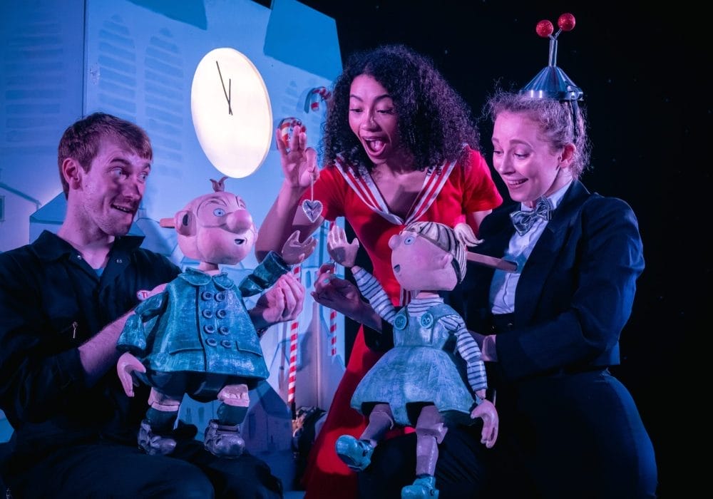 Three performers on stage interact with two large puppets. A man in black operates a puppet of a boy in a blue coat, while a woman in a suit and bow tie with a fun hat shaped like an antenna operates a puppet of a girl in a blue dress. Between them, a woman in a bright red dress holds up a shiny silver heart, smiling as the puppets reach toward it. A clock showing nearly midnight and candy canes decorate the background.